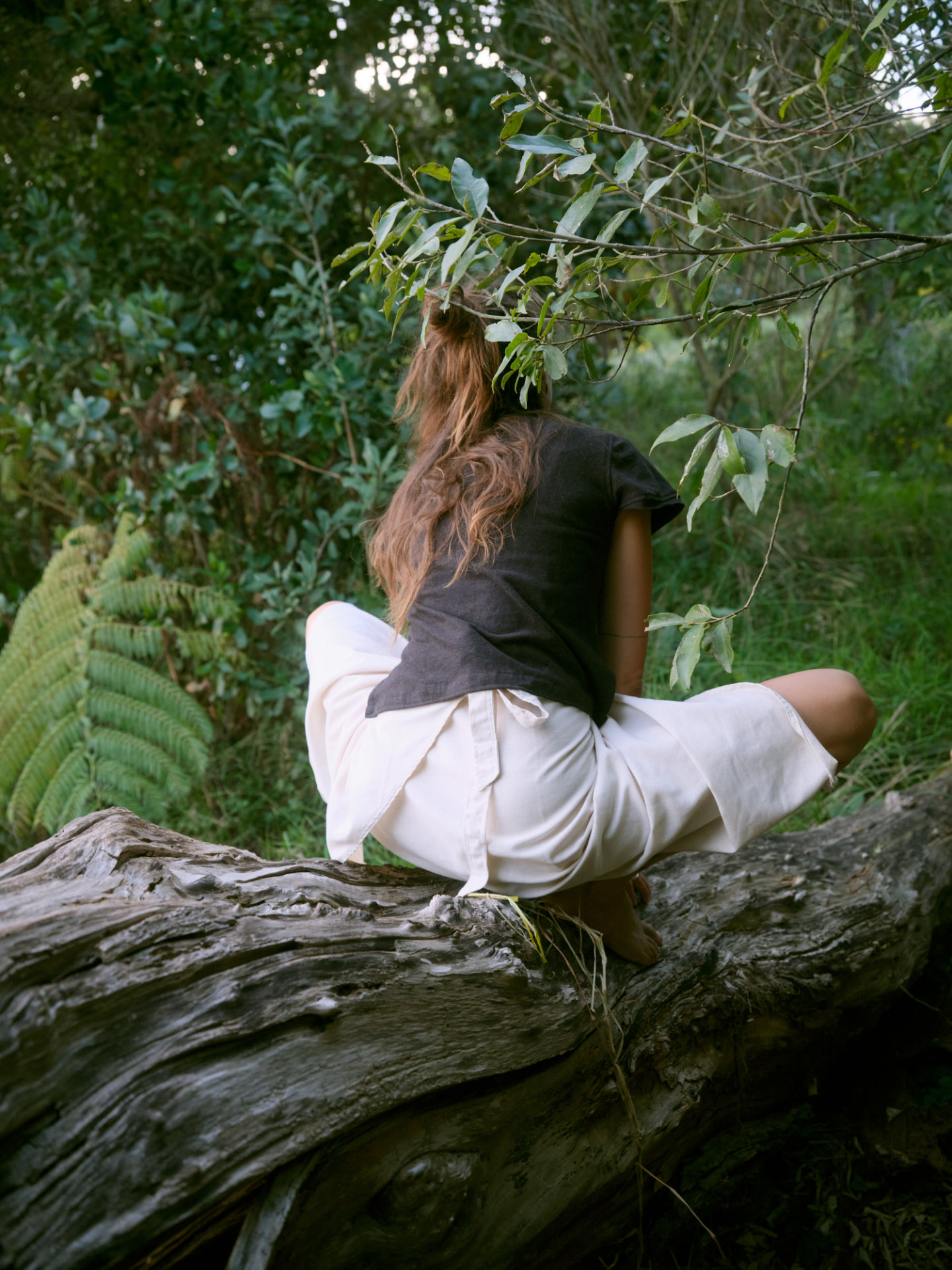 Woman in a dark tee and undyed wide trousers sitting on an old log among ferns