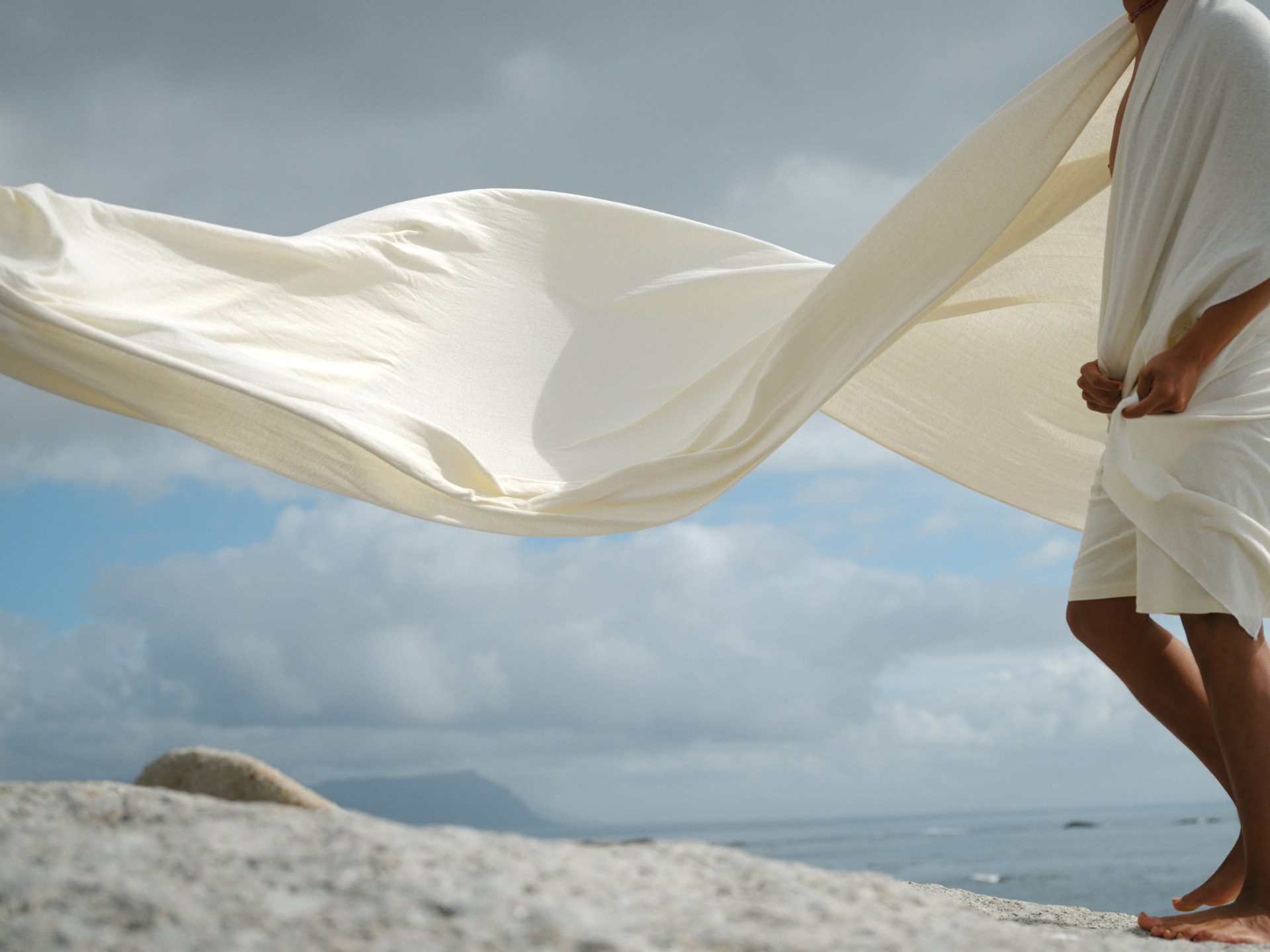 A figure on coastal granite holding undyed linen billowing in the sea wind